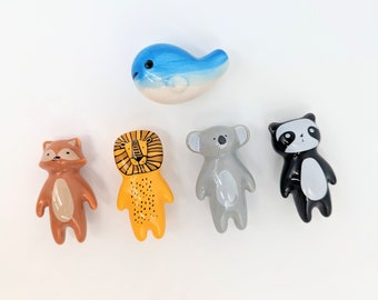 May include: Five ceramic animal figurines: a blue whale, a brown fox, a yellow lion, a gray koala, and a black and white panda. The figurines are all standing upright and have painted details.