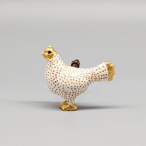 Pode incluir: Uma figurinha de frango de cerâmica branca com bolinhas douradas. O frango está em pé sobre suas patas com a cabeça inclinada para o lado. A figurinha é uma peça decorativa para a casa.
