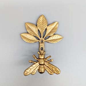 May include: Gold metal bee and leaf wall hook. The bee is in a flying position with its wings spread out. The leaves are arranged in a symmetrical pattern around the bee.