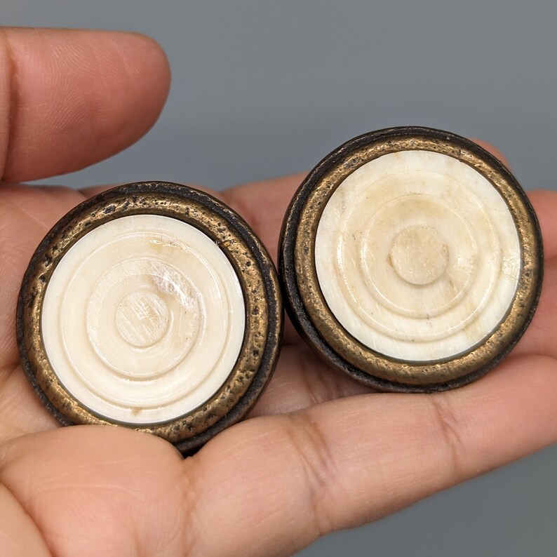 May include: Two round, decorative knobs with a brass finish and a white, concentric circle design.
