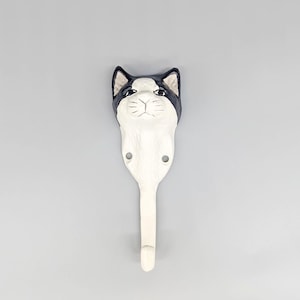 Puede incluir: Un gancho de pared blanco con forma de gato y detalles en negro en la cabeza y las orejas. El gancho tiene dos agujeros para el montaje y un solo gancho en la parte inferior. La cara del gato está detallada con ojos, nariz y bigotes.