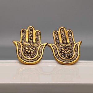 May include: A pair of gold-coloured earrings shaped like the Hamsa hand, a symbol of protection and good luck. The earrings are intricately detailed with floral patterns.