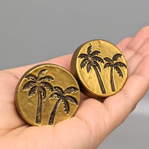 May include: Two round, brass-colored objects with a palm tree design. Each object has a dark brown palm tree illustration. The objects are held in a hand against a gray background.
