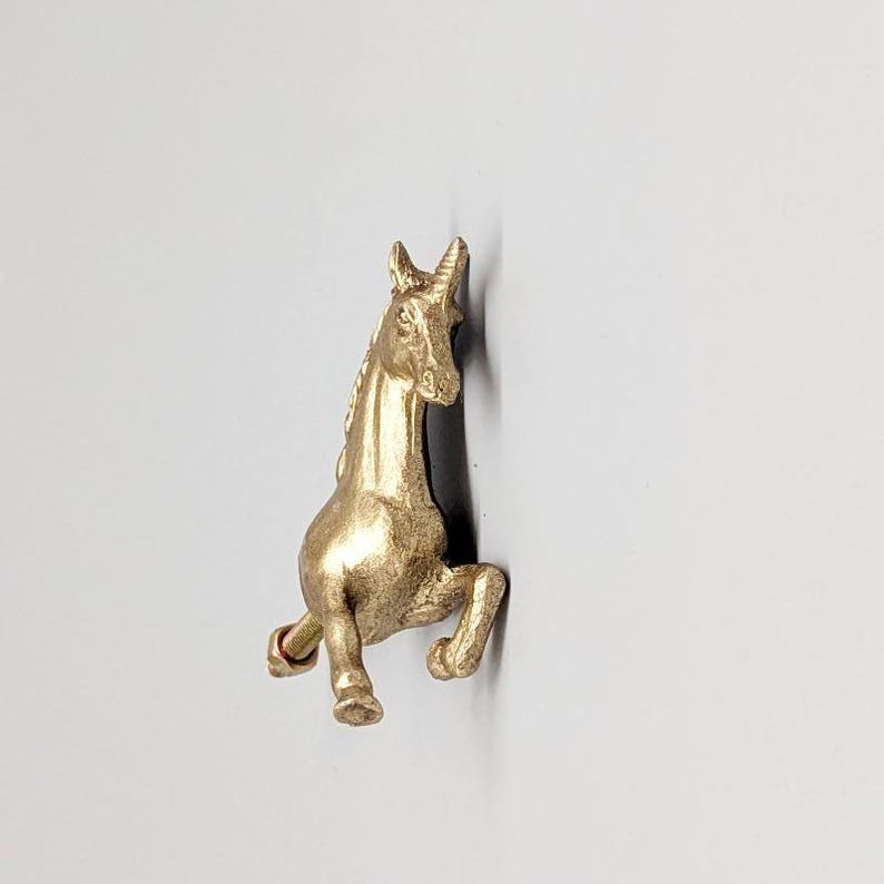 May include: A gold-colored unicorn figurine in a rearing pose. The sculpture has detailed features, including a flowing mane and a raised front leg. The unicorn is set against a plain background.