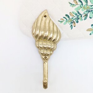 Könnte beinhalten: Ein goldfarbener Wandhaken in Muschelform. Der Haken hat ein detailliertes, strukturiertes Design und einen gebogenen Haken am unteren Ende. Der Haken ist aus Metall gefertigt und zur Wandmontage bestimmt. Der Haken ist ein dekorativer Artikel.