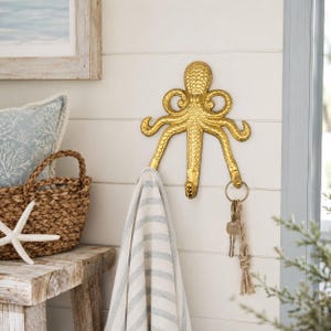 May include: A gold-colored wall hook shaped like an octopus with textured skin and multiple curved arms. The hook is mounted on a white paneled wall, with a striped fabric towel hanging from one arm and a set of metal keys hanging from another.
