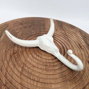 May include: White ceramic bull skull hook with a distressed finish. The hook is shaped like a bull's head with horns and a single hook on the bottom.