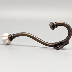 May include: A white ceramic knob with a fluted design is attached to a curved metal hook. The hook is finished in an antique brass colour.