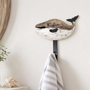 May include: A wall-mounted hook featuring a carved wooden whale figure with a weathered brown and white finish. The whale is attached to a vertical black metal hook, which holds a white and gray striped fabric towel hanging downward.