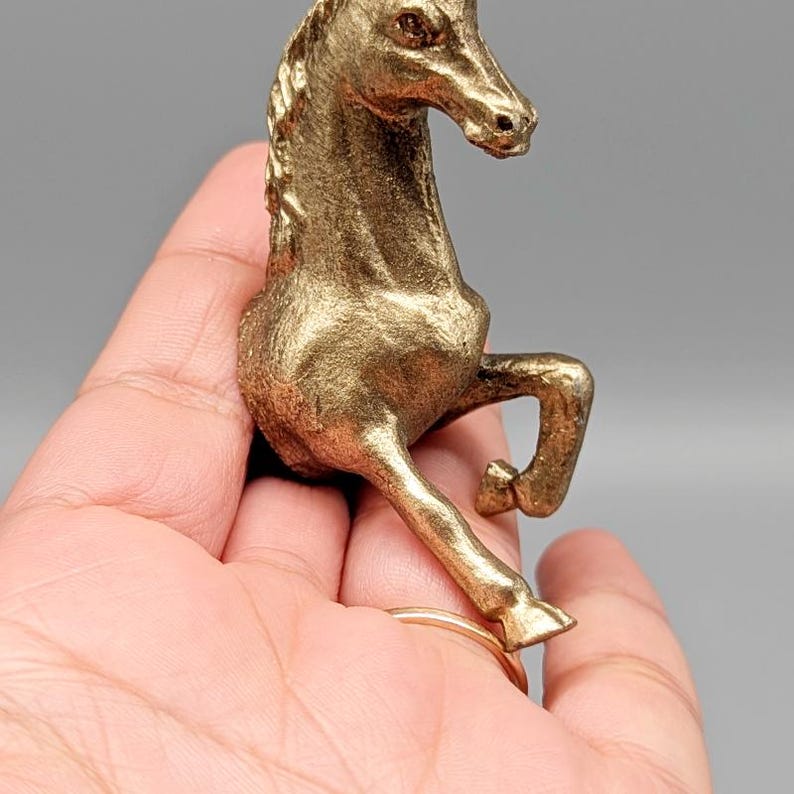 May include: A gold-colored horse figurine in a rearing pose. The sculpture has detailed features, including a flowing mane and a raised front leg. The horse is held in a hand against a gray background.