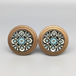 Puede incluir: Dos pomos de armario dorados con un diseño decorativo en blanco y negro con acentos azules. El diseño presenta un patrón circular con un círculo más pequeño en el centro.