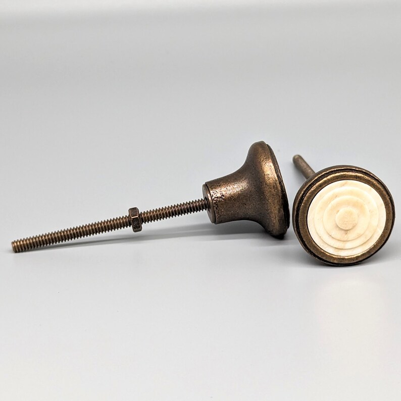 May include: Two antique bronze cabinet knobs with a white circular design. One knob has a threaded screw.