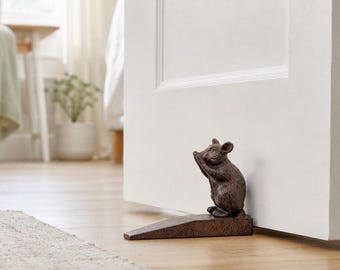 Op de afbeelding: Een donkerbruine, getextureerde metalen deurstopper in de vorm van een staande muis. De muis staat rechtop met de voorpootjes bij het gezicht en rust op een wigvormige basis die tegen de onderkant van een witte binnendeur is geplaatst.
