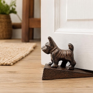 Pode incluir: Um batente de porta de ferro fundido castanho escuro em forma de um pequeno cão de pé com um acabamento texturizado. A figura do cão está montada numa base em forma de cunha, posicionada para manter uma porta branca aberta.