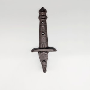 May include: A dark brown cast iron lighthouse-shaped bottle opener. The opener has a detailed design with a textured surface, featuring a lighthouse tower with circular windows and a flared base. The bottom has a pointed tip for leverage.