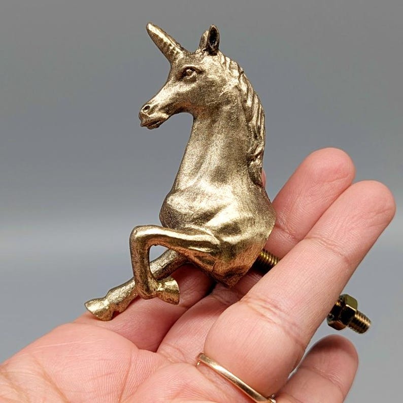 May include: A gold-colored unicorn figurine with a horn, mane, and legs. The unicorn is attached to a bolt and nut, suggesting it is a decorative hardware piece. The figurine is held in a hand against a gray background.
