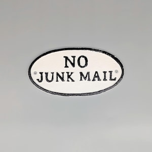 May include: An oval, white sign with the text "NO JUNK MAIL" in black, bold lettering. The sign has a black border and two small holes for fixing. A practical item for a home or office setting.