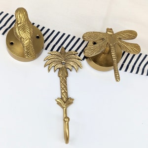 Puede incluir: Tres ganchos de metal dorado en forma de pájaro, libélula y palmera. Los ganchos están montados sobre un fondo blanco.
