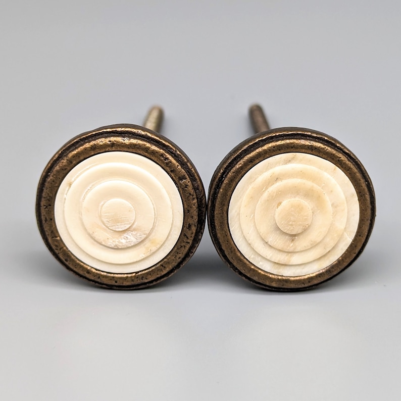 May include: Two round, white, concentric circle knobs with a brass finish. The knobs have a raised, textured design.