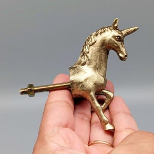 May include: A gold-colored unicorn-shaped cabinet knob. The unicorn is sculpted with detailed features, including a horn and flowing mane. A threaded bolt and nut are attached to the back for installation. The knob is held in a hand against a gray background.