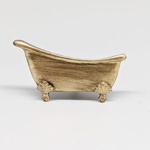 Könnte beinhalten: Eine kleine, goldfarbene Badewanne mit dekorativen fußförmigen Füßen. Die Wanne hat ein geschwungenes Design und eine strukturierte Oberfläche, die ihr ein Vintage-Aussehen verleiht. Die Gesamtästhetik ist elegant und dekorativ.