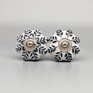 May include: Two white ceramic cabinet knobs with a floral design. Each knob has a black leaf pattern and a silver-colored center. The knobs are shaped like flowers and are set against a light gray background.