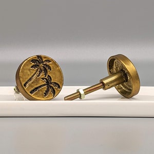 May include: Two brass cabinet knobs with a palm tree design. The knobs are round with a screw and nut for installation. The palm tree design is etched in black on the brass surface.