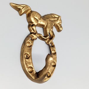 May include: A gold-coloured metal wall hook depicting a horse leaping over a horseshoe. The hook has a detailed, textured finish, and is suitable for hanging items. The design is a decorative accent for a rustic or equestrian-themed space.