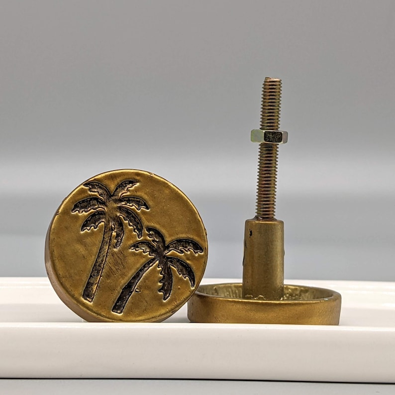 May include: A bronze-colored cabinet knob with a palm tree design. The knob is round with a raised image of two palm trees. The hardware includes a threaded rod, nut, and base for mounting. The knob is designed for furniture.