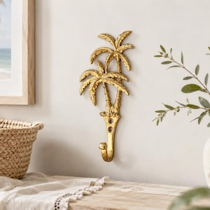 Op de afbeelding: Een goudkleurige metalen wandhaak met een decoratief ontwerp van twee palmbomen. De haak heeft een textuurafwerking en is voorzien van twee ronde montagegaten aan de basis van de stammen, met een enkele gebogen haak die naar beneden wijst.
