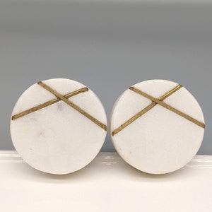 May include: Two round white marble drawer knobs with gold accents. The knobs have a simple, modern design with a criss-cross pattern.