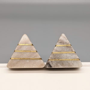 May include: Two small, triangular, decorative pyramids made of light grey marble with gold accents. The pyramids have three gold stripes on each side.