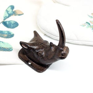 May include: A cast iron rhino head wall hook in a dark brown finish. The rhino head is detailed with textured skin and a prominent horn. The hook has a square base with a single hole for mounting. Decorative and functional.