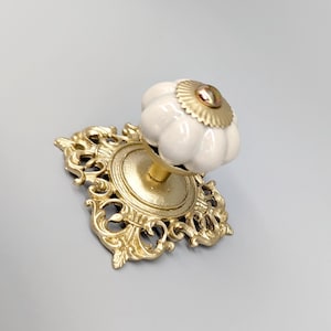 May include: A white ceramic knob with a gold base and ornate detailing. The knob has a scalloped edge and a gold accent in the center.