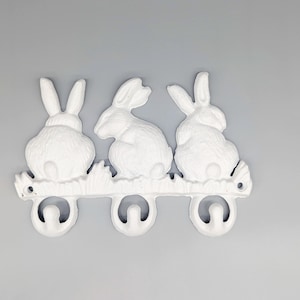 May include: White metal wall hook featuring three bunny rabbit figures. The rabbits are arranged horizontally and are connected to three curved hooks. The piece is painted white and is suitable for an Easter or spring theme.