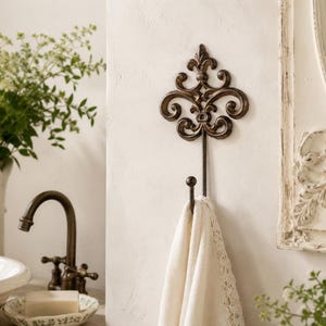 May include: A wall-mounted metal hook featuring an ornate, scrollwork design at the top with a dark, weathered bronze finish. A single, simple hook extends downward, currently holding a folded, cream-coloured fabric towel with lace trim.