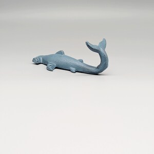 以下が含まれることがあります： イルカまたはクジラのような海洋動物の、水色の鋳造金属彫刻。彫刻は横向きに置かれ、尾は上向きに曲がっています。ひれや質感のある表面など、細部までこだわって作られています。