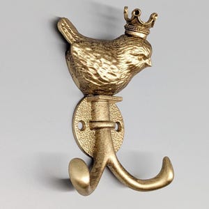 May include: A gold-coloured metal wall hook featuring a bird wearing a crown. The bird is perched above a round base with a curved hook below. The hook is designed for hanging items and adds a decorative touch to any space.