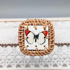 May include: A square-shaped, woven wicker coaster with a white tile featuring a red and white butterfly design.