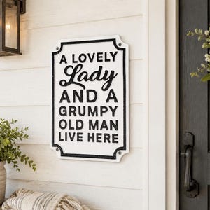 May include: A rectangular white metal wall sign with a black border and raised black lettering that reads A LOVELY Lady AND A GRUMPY OLD MAN LIVE HERE. The sign features rounded corners with small circular holes for mounting on a wall surface.