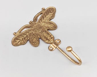 May include: A decorative brass-colored wall hook featuring a dragonfly design. The dragonfly has detailed wing patterns and is attached to a curved hook with a small ball at the end. The hook is designed for hanging items.