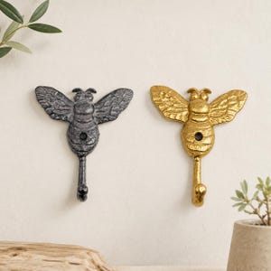 May include: Two wall hooks shaped like bees, one in a dark charcoal grey finish and one in a bright gold finish. Each bee features textured wings and a central body with a single circular mounting hole, extending into a curved hook at the base.