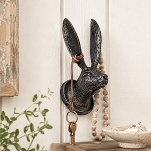 Puede incluir: Un gancho de pared de metal fundido, oscuro y texturizado, con forma de cabeza de liebre y orejas largas y erguidas. Un fino cordón de cuero marrón está atado a una oreja, sosteniendo una anilla de metal con dos llaves. La cabeza está en una base.