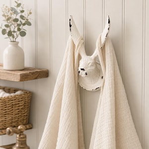 May include: A white wall-mounted hook shaped like a rabbit head with long ears, featuring a distressed finish with black accents. A cream-coloured textured fabric towel is draped over the rabbit ears, hanging down on both sides of the hook.