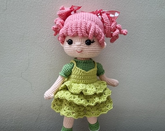 Könnte beinhalten: Handgemachte Häkelpuppe mit rosa Haaren, die in zwei lockigen Zöpfen mit roten Schleifen gehalten werden. Die Puppe trägt ein grünes Kleid mit einem geschichteten Rock und einem passenden grünen Oberteil. Die Puppe hat ein lächelndes Gesicht.