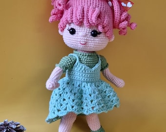 Könnte beinhalten: Handgemachte Häkelpuppe mit rosa Locken und einem grünen Kleid. Die Puppe hat einen hellen Hautton, rosige Wangen und schwarze Augen. Das Kleid hat einen gewellten Saum und wird mit grünen Schuhen kombiniert. Ein bezaubernder handgefertigter Artikel.