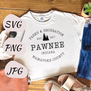 以下が含まれることがあります： 「Parks & Recreation Est. 1817 Pawnee Indiana Wapapoke County」と黒で書かれたグラフィックデザインが施された白いTシャツ。グラフィックデザインには松の木が2本描かれている。