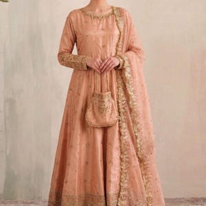 May include: A peach-colored floor-length gown with long sleeves, featuring intricate gold embroidery along the neckline, cuffs, and hem. The ensemble includes matching wide-leg trousers, a sheer dupatta with gold trim, and a small matching handbag.