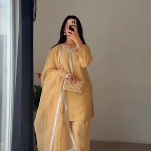 May include: A mustard-yellow salwar kameez ensemble. The outfit includes a tunic, pants, and a sheer dupatta with pearl accents. A matching clutch is held. The outfit is worn with sandals. The outfit is reflected in a mirror.