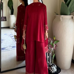 May include: A deep red long-sleeved tunic and matching wide-leg trousers set. The garment features intricate tonal embroidery and embellishments along the sleeve cuffs and the hem of the trousers. A coordinating red scarf is draped over the shoulders.